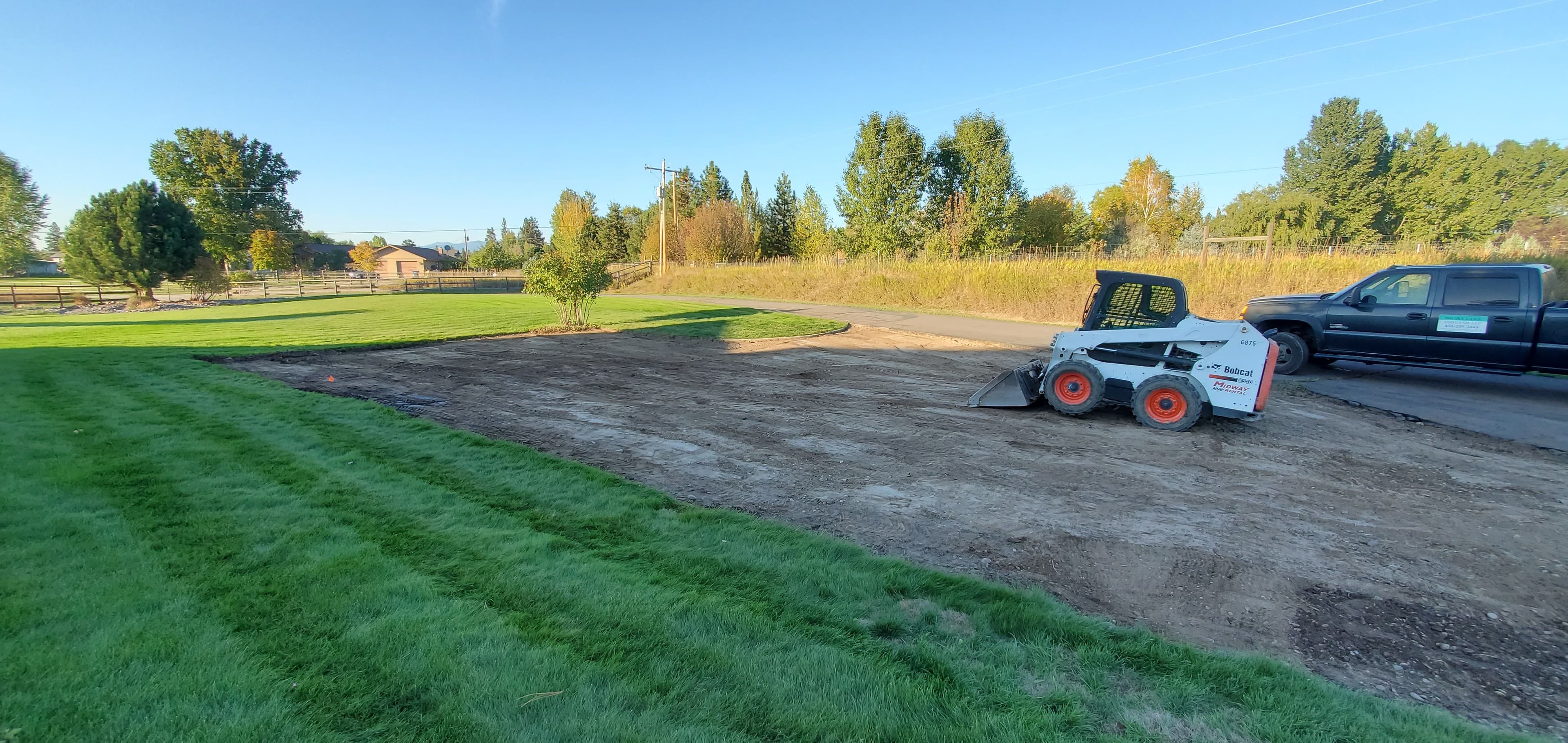 Bobcat loader on dirt, preparing landscaping near a grassy area and trees in a residential setting.