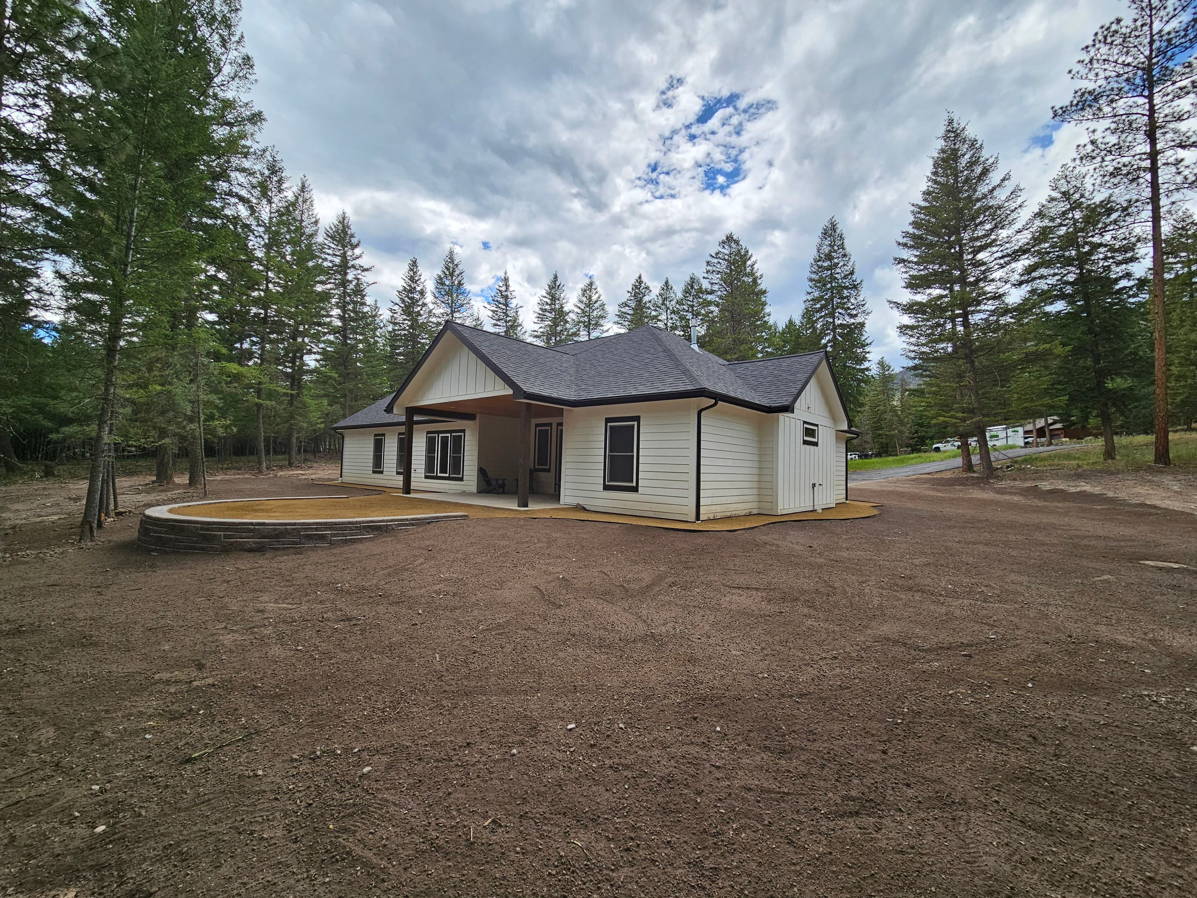 Modern white house surrounded by pine trees and gravel yard under a cloudy sky.