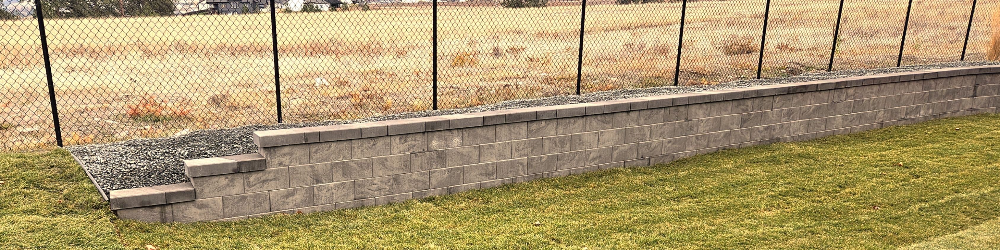 Retaining wall with gravel top, surrounded by fence and grassy field in background.