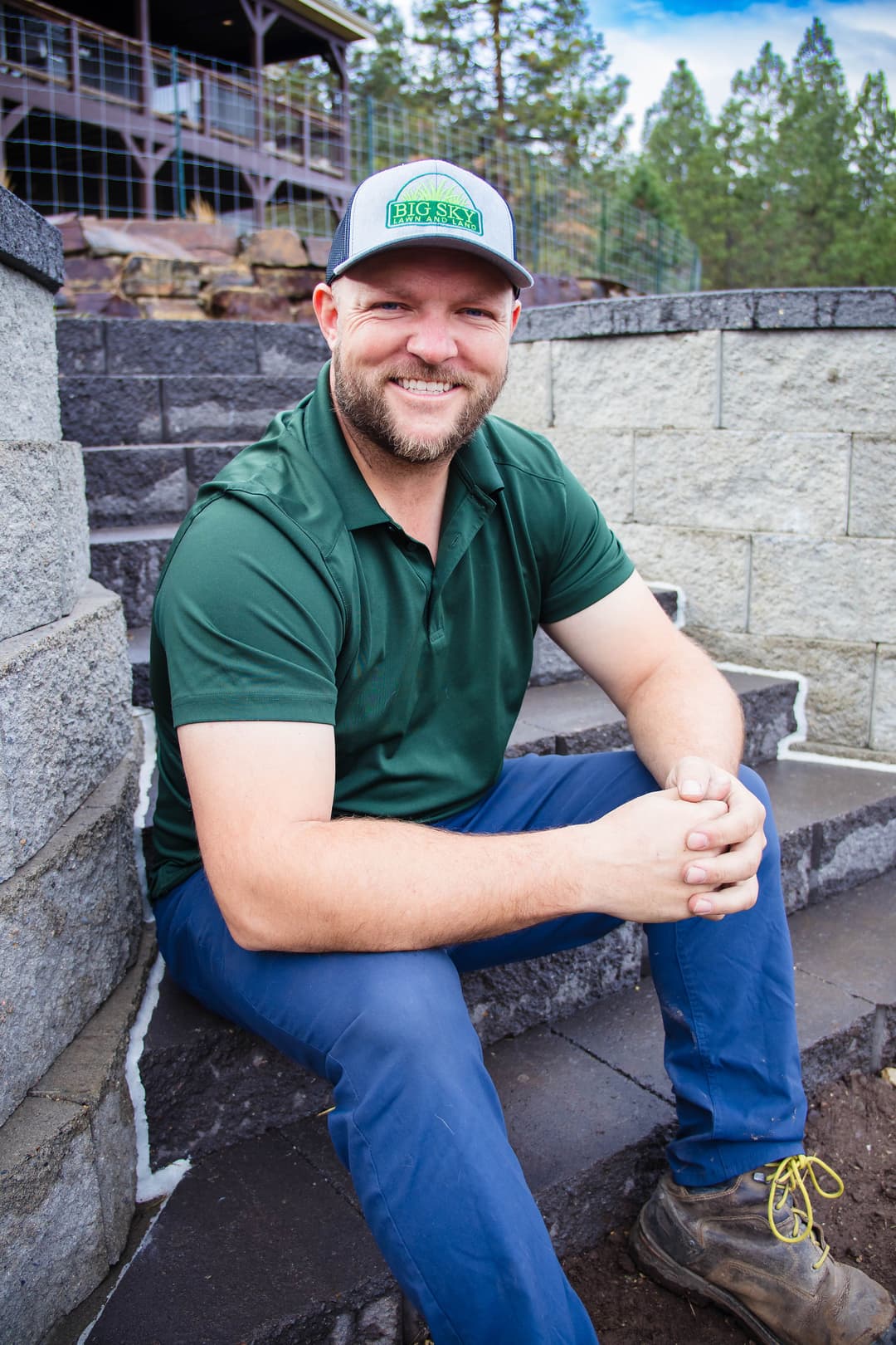 Smiling man in green shirt and hat sitting on outdoor stone stairs, surrounded by trees.