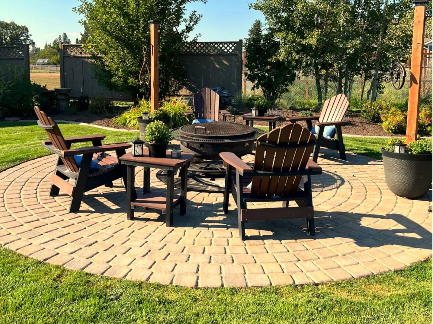 Cozy outdoor fire pit area with wooden chairs and plants on a brick patio in a garden setting.