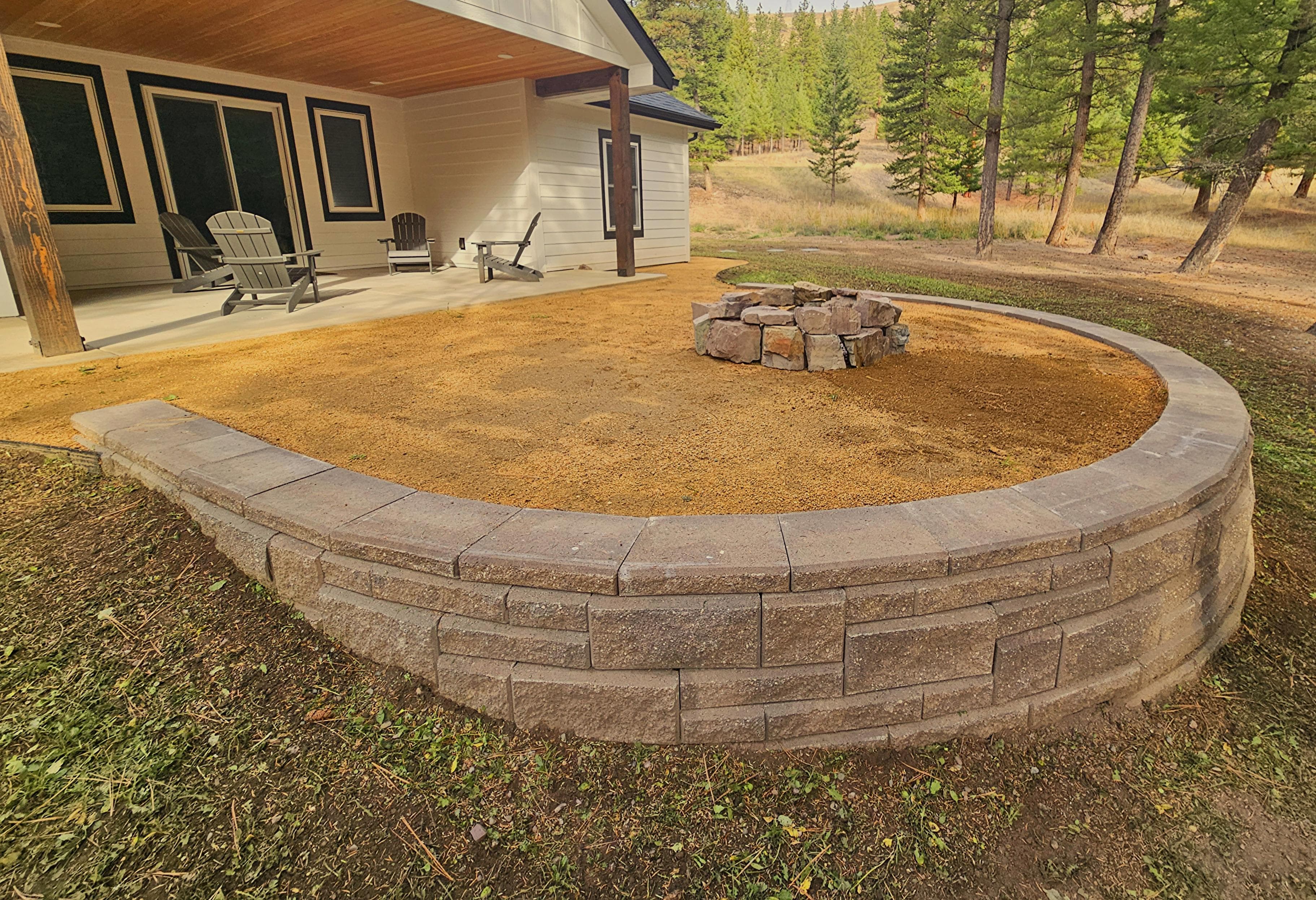Stone fire pit area with a curved retaining wall, surrounded by dirt and trees in a scenic setting.