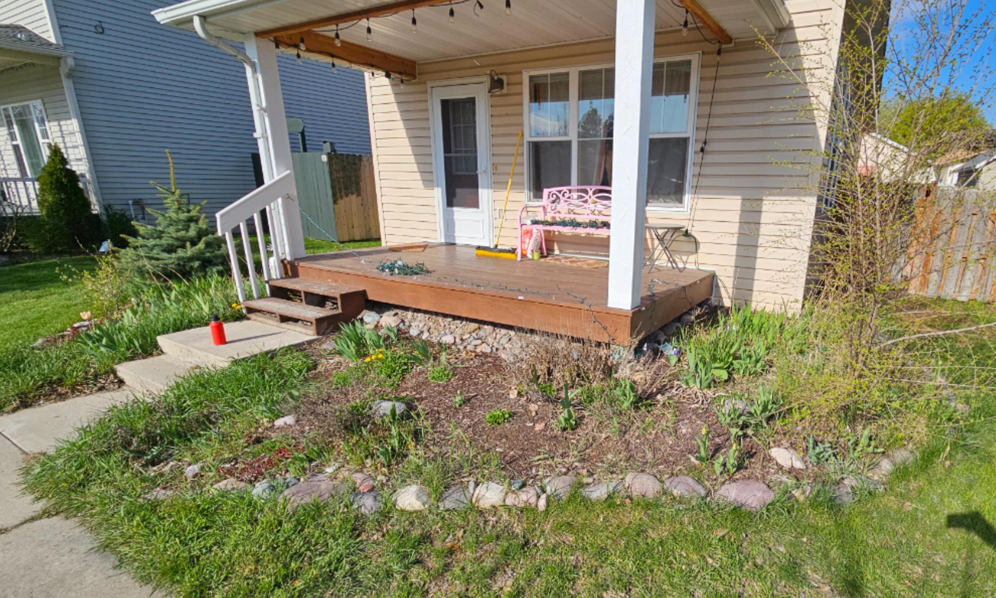 Front porch of a house with wooden steps, garden, and seating area on a sunny day.