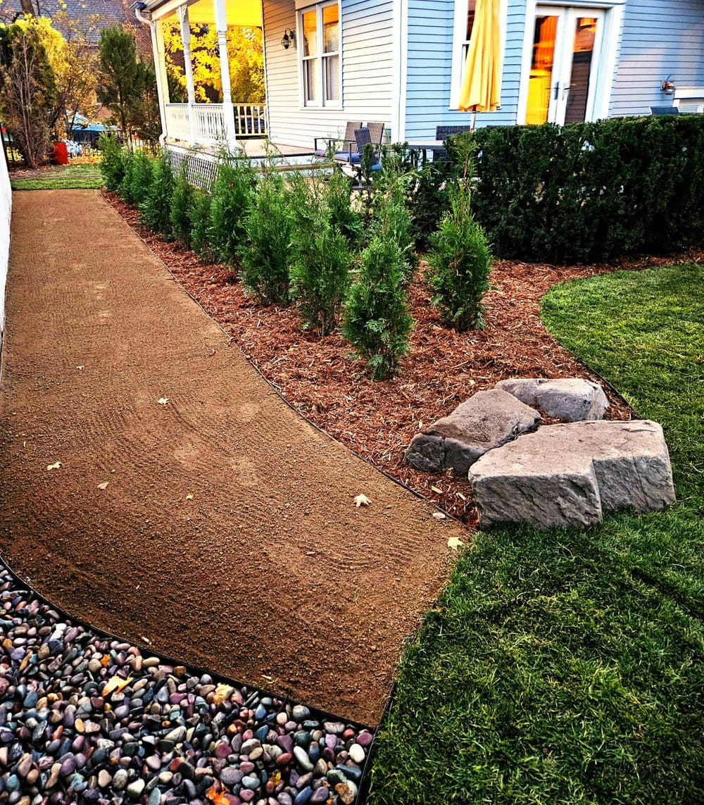 Curved garden pathway with mulch, shrubs, and decorative stones beside a charming blue house.
