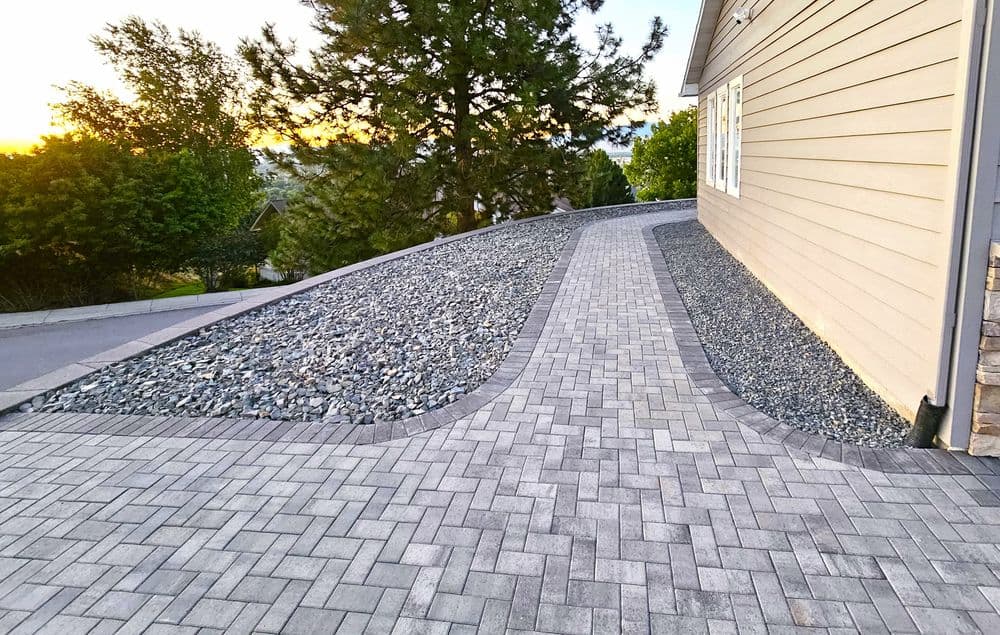 Paved pathway with landscape stones beside a house, sunset visible in the background.
