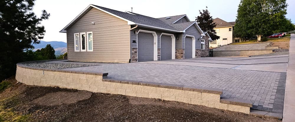 Modern house with gray siding, garage, and stone wall, set on a hillside with a paved driveway.
