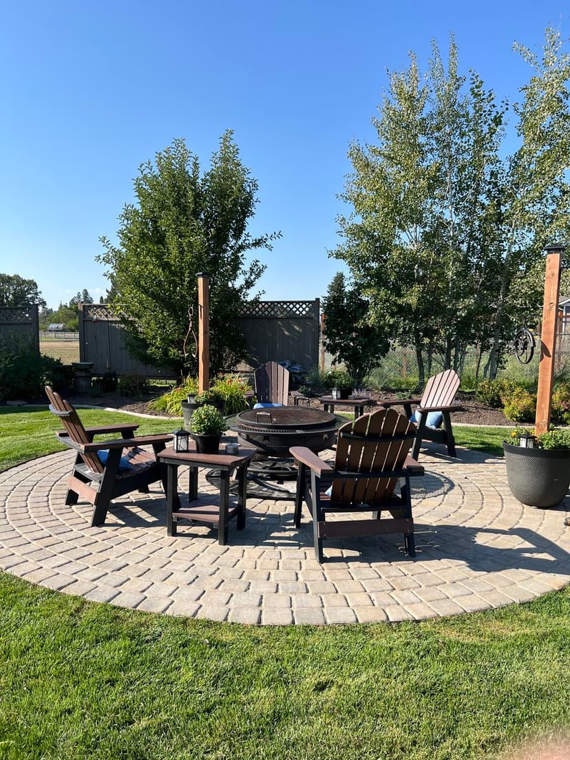 Outdoor fire pit area with Adler chairs, surrounded by landscaped greenery and blue sky.