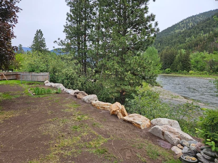 Riverbank landscape featuring stone border, greenery, and distant mountains under a cloudy sky.
